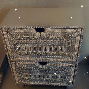 Puede incluir: Una pequeña cómoda de dos cajones con un acabado gris oscuro. Los cajones están decorados con un mosaico de pequeños diamantes de imitación iridiscentes.