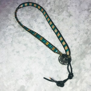 May include: A teal, gold, and cream beaded bracelet with a black leather cord and a round metal clasp.