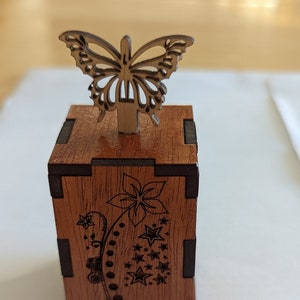 May include: A wooden box with a laser-cut butterfly on top. The box is brown with dark brown accents and features a floral and star design. The butterfly is intricately cut, adding a decorative touch to this small wooden box.