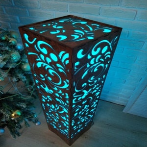 May include: A tall, wooden floor lamp with a square base and intricate cut-out designs. The lamp is illuminated with a bright blue light, creating a decorative and modern look.