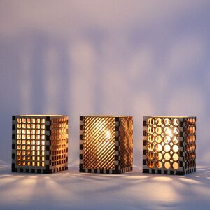 Może przedstawiać: Trzy drewniane świeczniki z różnymi wzorami geometrycznymi. Każdy świecznik ma w środku świecę, która rzuca ciepłe światło przez wycięcia.