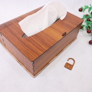 Puede incluir: Un porta pañuelos de madera con un patrón de veta de madera y una pequeña cerradura en el lateral. La caja está abierta y se puede ver un pañuelo blanco en el interior.