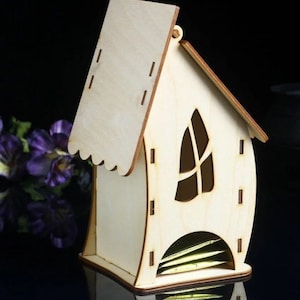 Puede incluir: Un soporte de bolsitas de té de madera con forma de casa, con una ventana recortada y una ranura para las bolsitas de té. El soporte está hecho de madera clara y tiene un acabado blanco.
