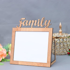 以下が含まれることがあります： 「Family」という言葉が上に彫られた木製の写真フレーム。フレームは空で、写真を入れる準備ができています。