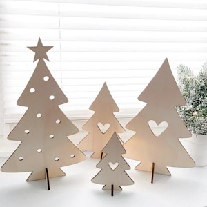 Op de afbeelding: Vier houten kerstboomversieringen van verschillende formaten. De grootste boom heeft een ster bovenop en cirkelvormige uitsparingen. De andere bomen hebben hartvormige uitsparingen. Alle zijn gemaakt van lichtgekleurd hout en staan rechtop.
