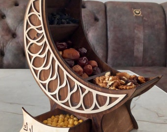 May include: A wooden crescent moon-shaped snack bowl with three compartments and a drawer. The bowl is decorated with intricate cutouts and the words "Ramadan Mubarak" are inscribed on the drawer. The bowl is filled with dried fruit and nuts.