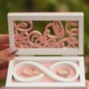 May include: White wooden ring box with a pink infinity symbol design inside. The box has a clear window on the top and a pink floral design on the inside of the lid.