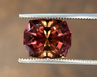 Op de afbeelding: Een gefacetteerde edelsteen die door een pincet wordt vastgehouden. De edelsteen heeft een mix van rode, roze en gouden tinten. De slijpvorm is een gemodificeerde achthoekige vorm, met meerdere facetten die licht reflecteren. De pincet is zilverkleurig.