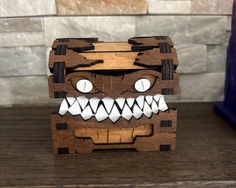 May include: A wooden box with a monster face design. The box is made of dark brown wood with a light brown top. The monster face has white teeth and white oval eyes. The box is sitting on a dark brown wooden surface.