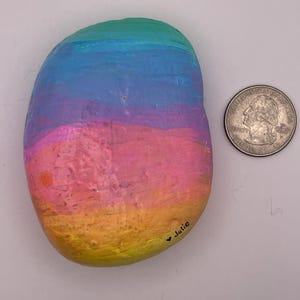 May include: A smooth, oval-shaped river rock painted with horizontal bands of gradient colors including yellow, orange, pink, purple, and blue. The name Julie is written in black ink near the bottom edge next to a US quarter coin for scale.