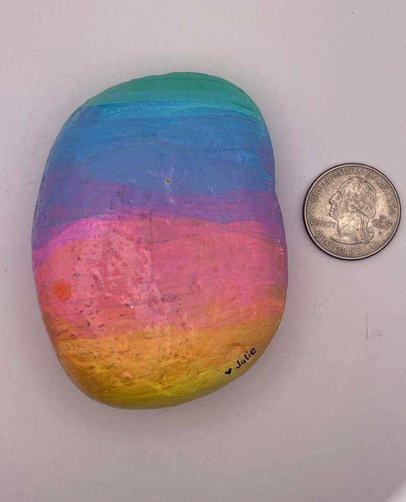 May include: A smooth, oval-shaped river rock painted with horizontal bands of gradient colors including yellow, orange, pink, purple, and blue. The name Julie is written in black ink near the bottom edge next to a US quarter coin for scale.