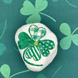 Op de afbeelding: Een wit beschilderde steen met een groen klaverbladontwerp. Het klaverblad heeft een geruit hart, gestreepte bladeren en gestippelde details. De achtergrond is een groene stof met klaverbladpatronen.
