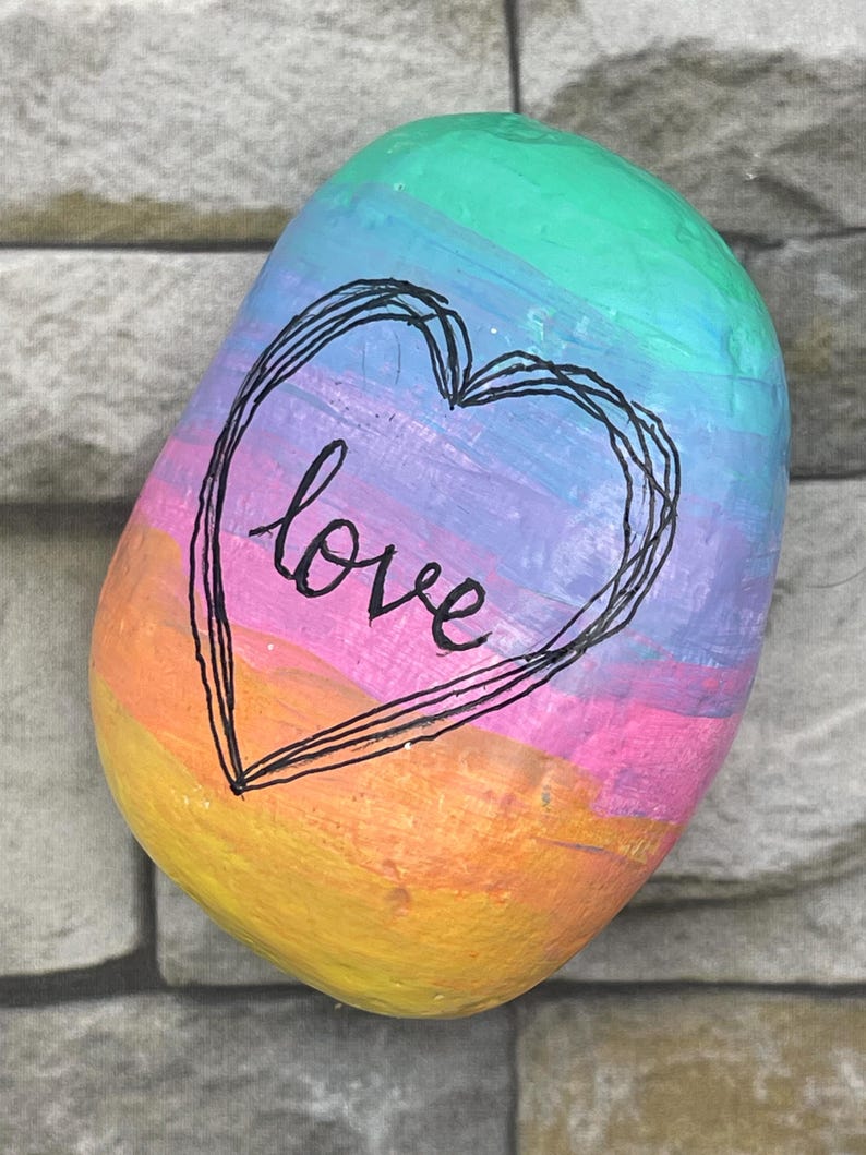 May include: A smooth, oval-shaped river rock painted with horizontal stripes in a rainbow gradient of yellow, orange, pink, purple, blue, and green. A black hand-drawn heart outline surrounds the lowercase word love written in black script.