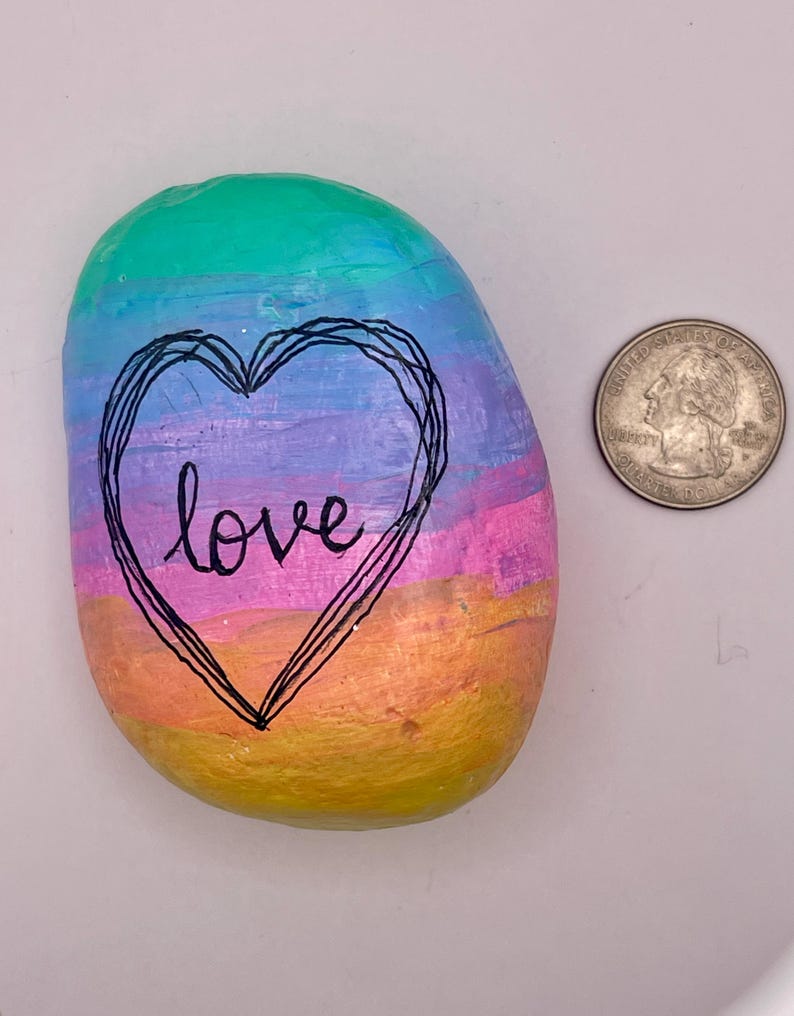 May include: A smooth, oval-shaped river rock painted with horizontal bands of teal, blue, purple, pink, orange, and yellow. A black hand-drawn heart outline surrounds the word love in cursive script. A US quarter is shown for scale.