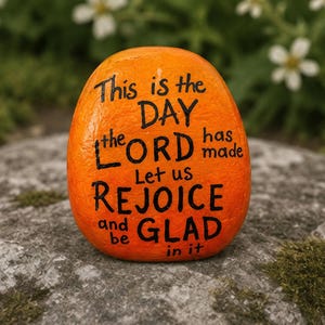 May include: A smooth, rounded orange stone featuring black hand-painted text that reads: This is the DAY the LORD has made Let us REJOICE and be GLAD in it. The stone has a vibrant, solid orange surface with clear, bold black lettering.