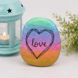 May include: A smooth, rounded river stone painted with horizontal stripes in shades of teal, blue, purple, pink, orange, and yellow. A black hand-drawn heart outline surrounds the word love written in black cursive script on the center.