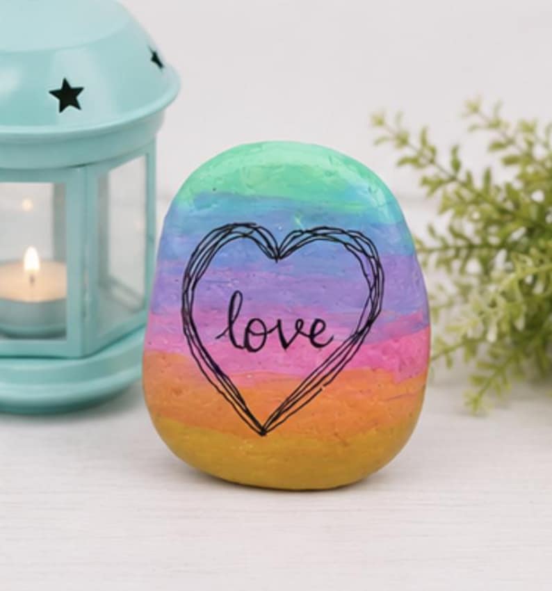 May include: A smooth, rounded river stone painted with horizontal stripes in shades of teal, blue, purple, pink, orange, and yellow. A black hand-drawn heart outline surrounds the word love written in black cursive script on the center.