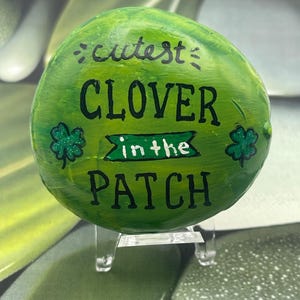 Op de afbeelding: Een handgeschilderde groene steen met de woorden "cutest CLOVER in the PATCH" in zwarte letters. De steen heeft twee glitter klaverontwerpen. De steen wordt op een transparante standaard getoond.