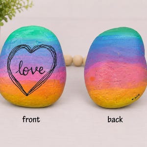 May include: Two smooth, rounded stones painted with a vibrant rainbow gradient of green, blue, purple, pink, orange, and yellow. The front stone features a black hand-drawn heart outline with the word love inside. The back stone has the name Julie.