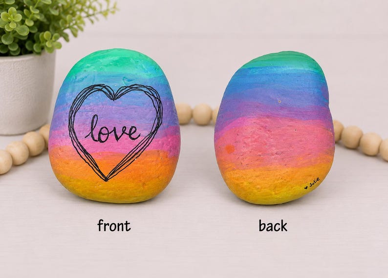 May include: Two smooth, rounded stones painted with a vibrant rainbow gradient of green, blue, purple, pink, orange, and yellow. The front stone features a black hand-drawn heart outline with the word love inside. The back stone has the name Julie.
