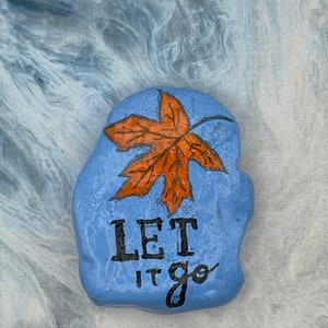 Puede incluir: Una piedra pintada a mano, de color azul claro, con un diseño de hoja naranja y las palabras "LET IT go" en negro. La piedra tiene una superficie texturizada, probablemente un objeto decorativo o pisapapeles.