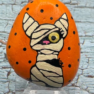 Gato momia con ojos que brillan en la oscuridad, arte de Halloween, piedra pintada, pintura en piedra