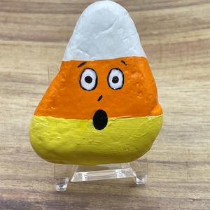 Può includere: Un sasso dipinto a forma di caramella di mais. Il sasso è diviso in sezioni bianche, arancioni e gialle e presenta un volto sorpreso con grandi occhi e bocca aperta. È esposto su un supporto di plastica trasparente.