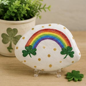 Op de afbeelding: Een wit beschilderde steen met een kleurrijke regenboog die tussen twee groene klaverbladen boogt. Het oppervlak van de steen is versierd met kleine gouden stippen. De steen staat op een doorzichtige plastic standaard met een los klaverblad.