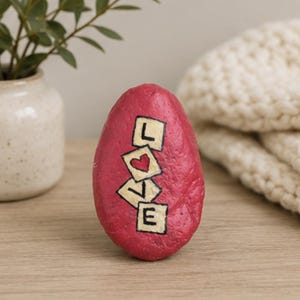 Op de afbeelding: Een gladde, ovaalvormige steen die in een levendige roze tint is geschilderd. Het oppervlak bevat vier vierkante tegels die verticaal zijn gerangschikt, elk met een zwarte letter die het woord LOVE vormt, met een rood hartje in plaats van de O.