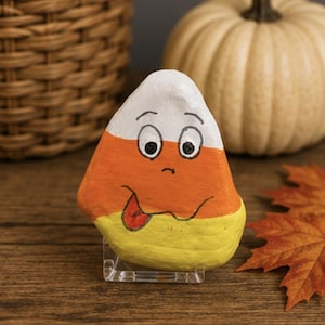 May include: A triangular rock painted to resemble a piece of candy corn with horizontal bands of white, orange, and yellow. The rock features a hand-painted cartoon face with wide eyes, a small nose, and a mouth with a red tongue sticking out.
