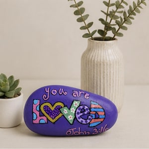 Pode incluir: Uma pedra oval e lisa de cor roxa, pintada com as palavras "you are loved" em letras de forma coloridas e estampadas. A letra "o" é substituída por um coração amarelo e o texto "John 3:16" está escrito em rosa na parte inferior.