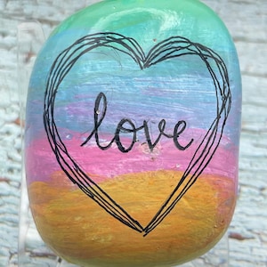 May include: A smooth, rounded river stone painted with horizontal bands of green, blue, purple, pink, and orange. A black hand-drawn heart outline surrounds the lowercase word love written in black script in the center of the stone.