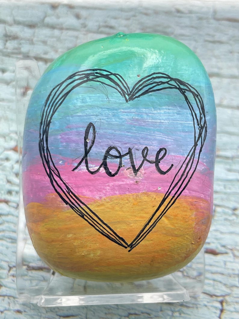 May include: A smooth, rounded river stone painted with horizontal bands of green, blue, purple, pink, and orange. A black hand-drawn heart outline surrounds the lowercase word love written in black script in the center of the stone.