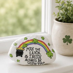 May include: A white painted rock featuring a hand-painted black pot of gold, a colorful rainbow, and green shamrocks. The text reads, "May the luck of the Irish always be with you." The rock is positioned next to a speckled cream-colored planter.