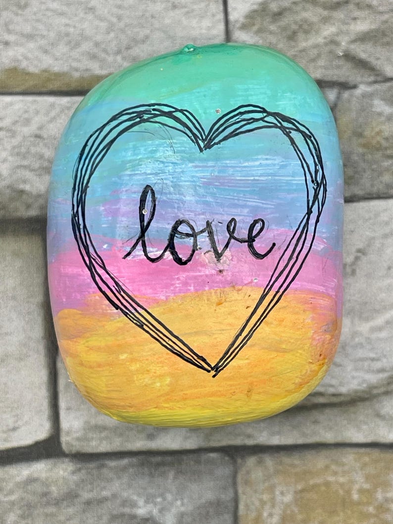 May include: A smooth, rounded river stone painted with horizontal bands of yellow, pink, light blue, and green. A black hand-drawn heart outline surrounds the word love, which is written in black cursive script across the center of the stone.