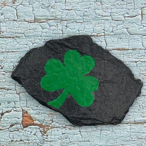 Könnte beinhalten: Ein unregelmäßig geformter schwarzer Schieferstein mit einem hellgrünen Kleeblatt darauf. Das Kleeblatt hat drei Blätter und ist auf dem Stein zentriert. Der Hintergrund ist hellblaues, verwittertes Holz.