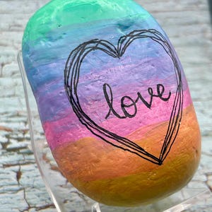 May include: A smooth, oval-shaped river rock painted with horizontal bands of rainbow colors including green, blue, purple, pink, orange, and yellow. A black hand-drawn heart outline surrounds the cursive word love written in black ink.