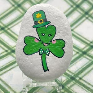 May include: A hand-painted rock with a green shamrock design. The shamrock has a cute face and is wearing a green hat with a gold buckle and a green bow tie.