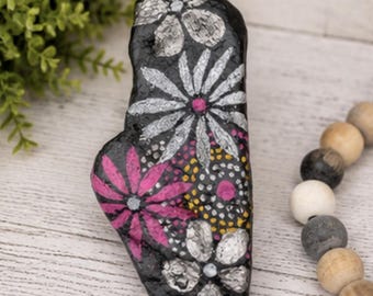 Puede incluir: Una piedra negra de forma irregular pintada con un diseño floral. La obra presenta pétalos plateados y rosas, con pequeños puntos amarillos y blancos que rellenan los espacios entre las flores. La superficie tiene un acabado brillante y texturizado.