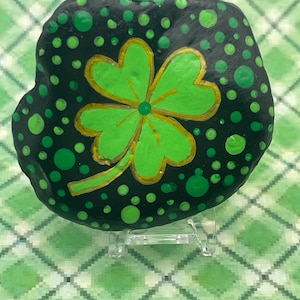 May include: A hand-painted, black rock featuring a green four-leaf clover with gold accents. The rock is decorated with various sized green dots. The clover is centered on the rock, and the design is displayed on a clear stand.