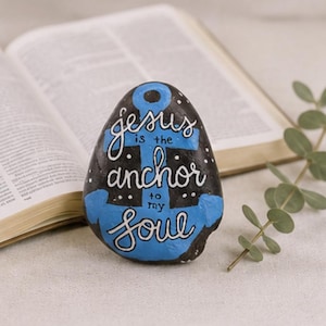 May include: A smooth, rounded black stone painted with a bright blue anchor design. The white hand-lettered text reads, "Jesus is the anchor to my soul," accented with small white dots resembling stars against the dark background of the stone.