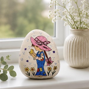 May include: A smooth, cream-colored stone featuring a hand-painted illustration of a figure wearing a pink hat and blue overalls, holding a small yellow bird. The design includes small yellow flowers and blue dots on a light background.