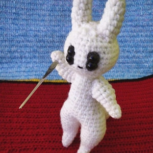 May include: White crocheted bunny rabbit toy with black eyes. The bunny is holding a silver crochet hook.