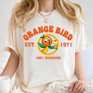 A woman wearing a white t-shirt with an orange bird graphic and text, sitting on a bed with a white blanket.
