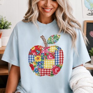 Op de afbeelding: Een lichtblauw t-shirt met korte mouwen en een grote, centrale appelafbeelding. De appel is samengesteld uit verschillende kleurrijke patchwork-stofpatronen, waaronder bloemenprints, geel gingham en rood-blauwe ruitjes.