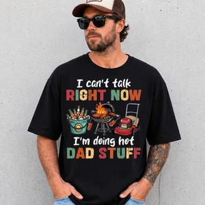 May include: Black t-shirt with the text "I can't talk RIGHT NOW I'm doing hot DAD STUFF". The graphic includes a grill, a lawnmower, and a bucket of beer. The shirt is a casual wear item.
