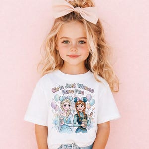 Könnte beinhalten: Ein junges Mädchen trägt ein weißes T-Shirt mit dem Text "Girls Just Wanna Have Fun". Das Shirt zeigt eine farbenfrohe Illustration von zwei Zeichentrickfiguren mit Luftballons und Getränken. Das Mädchen hat blondes Haar und eine große rosa Schleife.