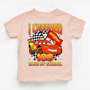 A pink t-shirt with a graphic design featuring a cartoon car and the text "I CRUSHED 100 DAYS OF SCHOOL".