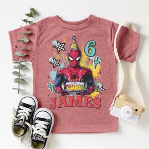 A pink t-shirt with a Spiderman graphic and the number 6, along with a pair of sneakers and a camera, all arranged on a white background.