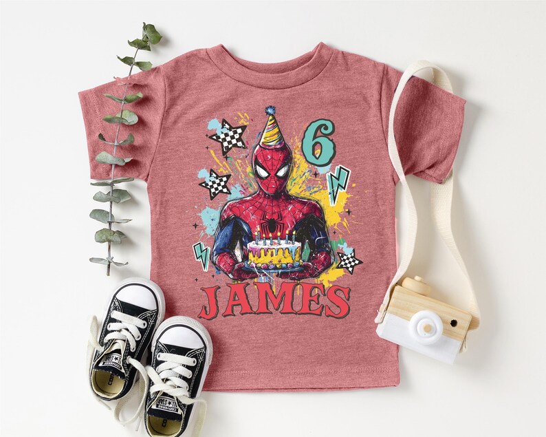 A pink t-shirt with a Spiderman graphic and the number 6, along with a pair of sneakers and a camera, all arranged on a white background.